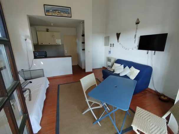 Lou cigaloun, proche plage, parking, terrasse Salon lumineux avec canapé bleu, table pliante et kitchenette. Mur blanc.