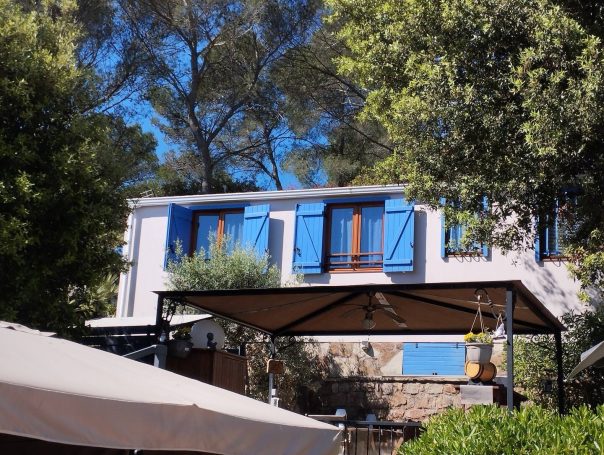 Maison blanche avec volets bleus, entourée d'arbres, terrasse à l'avant.
