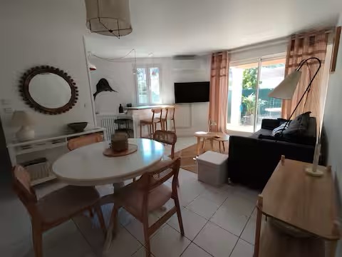 T2 climatisé avec jardin à 5' de la plage Intérieur d'un salon moderne avec table ronde, chaises, canapé et fenêtres éclairantes.
