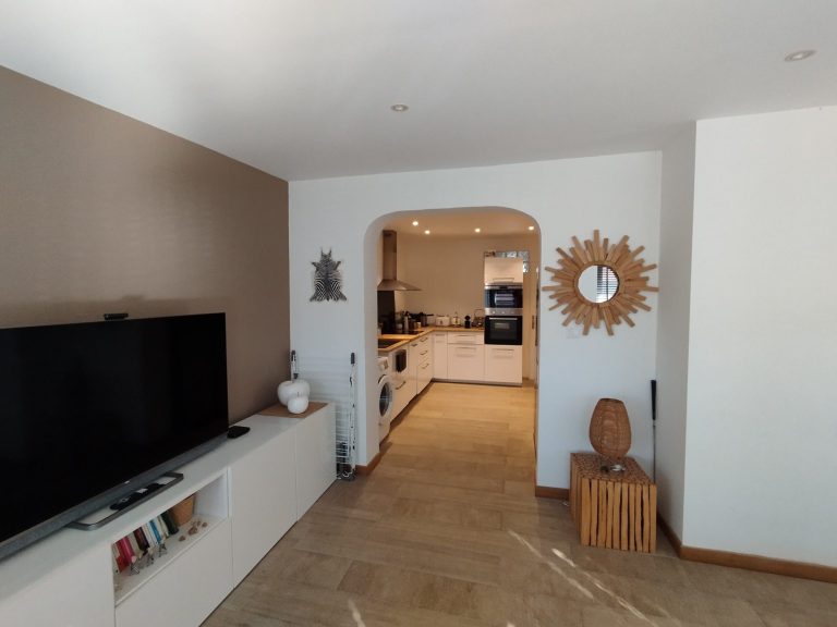 Salon moderne avec télévision, miroir soleil et une cuisine lumineuse.