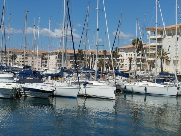 Port de plaisance avec plusieurs voiliers amarrés et bâtiments en arrière-plan.